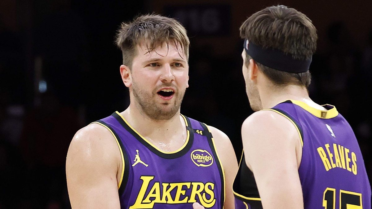 Luka Doncic speaks to Austin Reaves, whose back faces the camera, in purple Lakers uniforms.