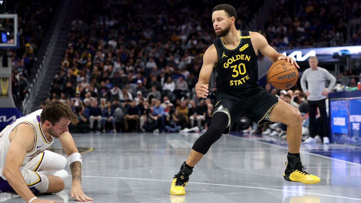 Stephen Curry, in a black Warriors No. 30 jersey, dribbles as Luka Doncic, in a white Lakers jersey, is down on the floor.