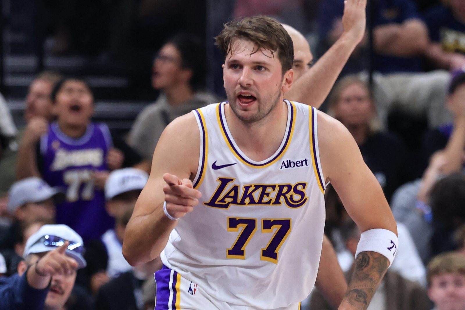 Luka Doncic, in a white Lakers No. 77 jersey, points and shouts on the court with fans behind him.